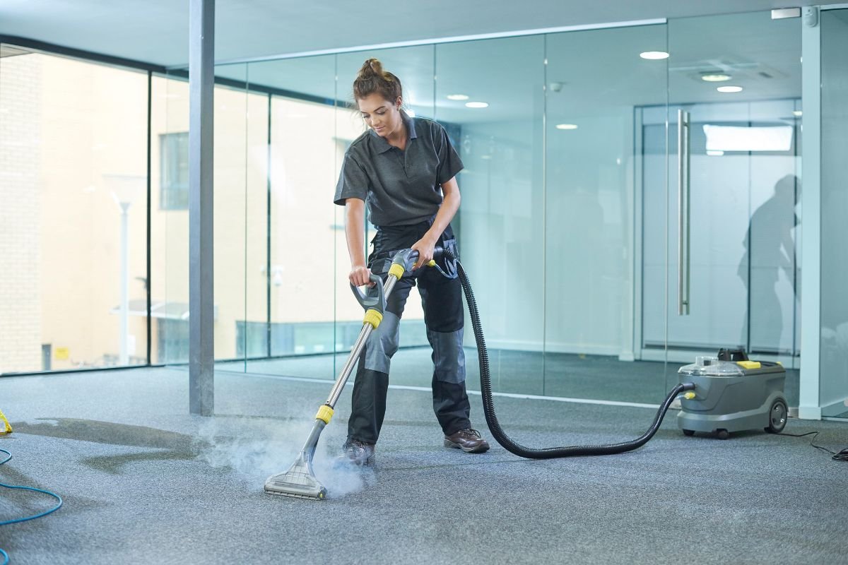 A person in a dark polo shirt cleans a carpet with a steam cleaner, surrounded by glass walls in a modern office space.