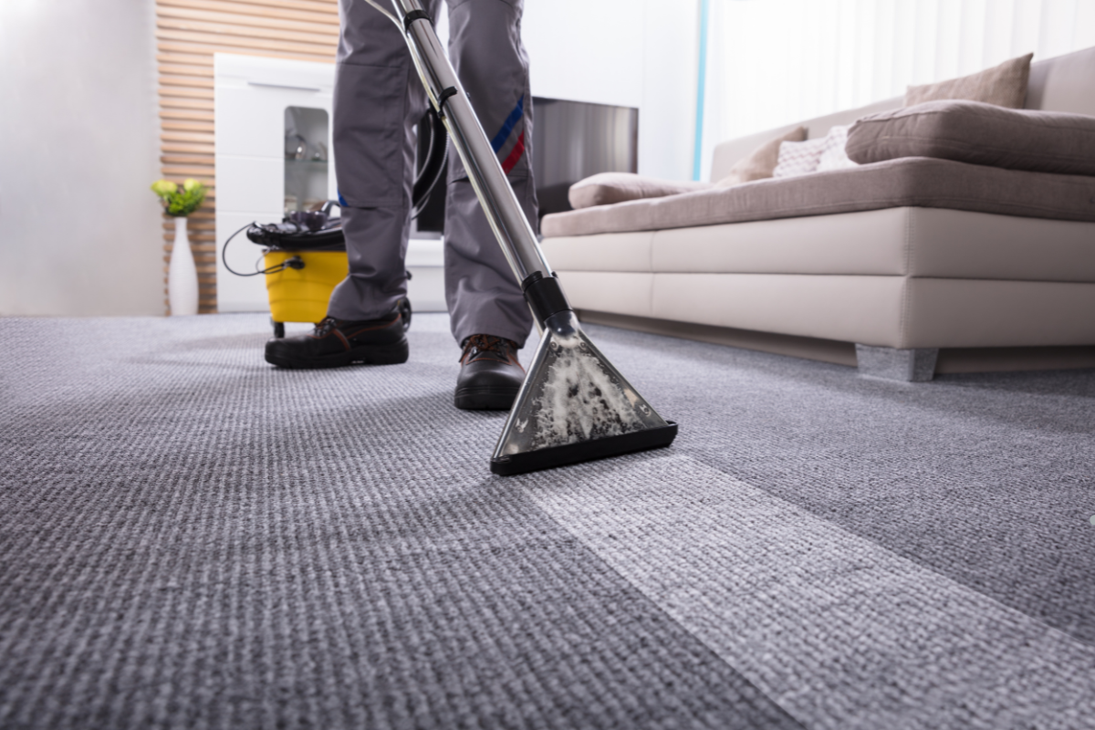 A person in work attire cleans a gray carpet with a professional extractor, a yellow bucket nearby and a modern living room in the background.